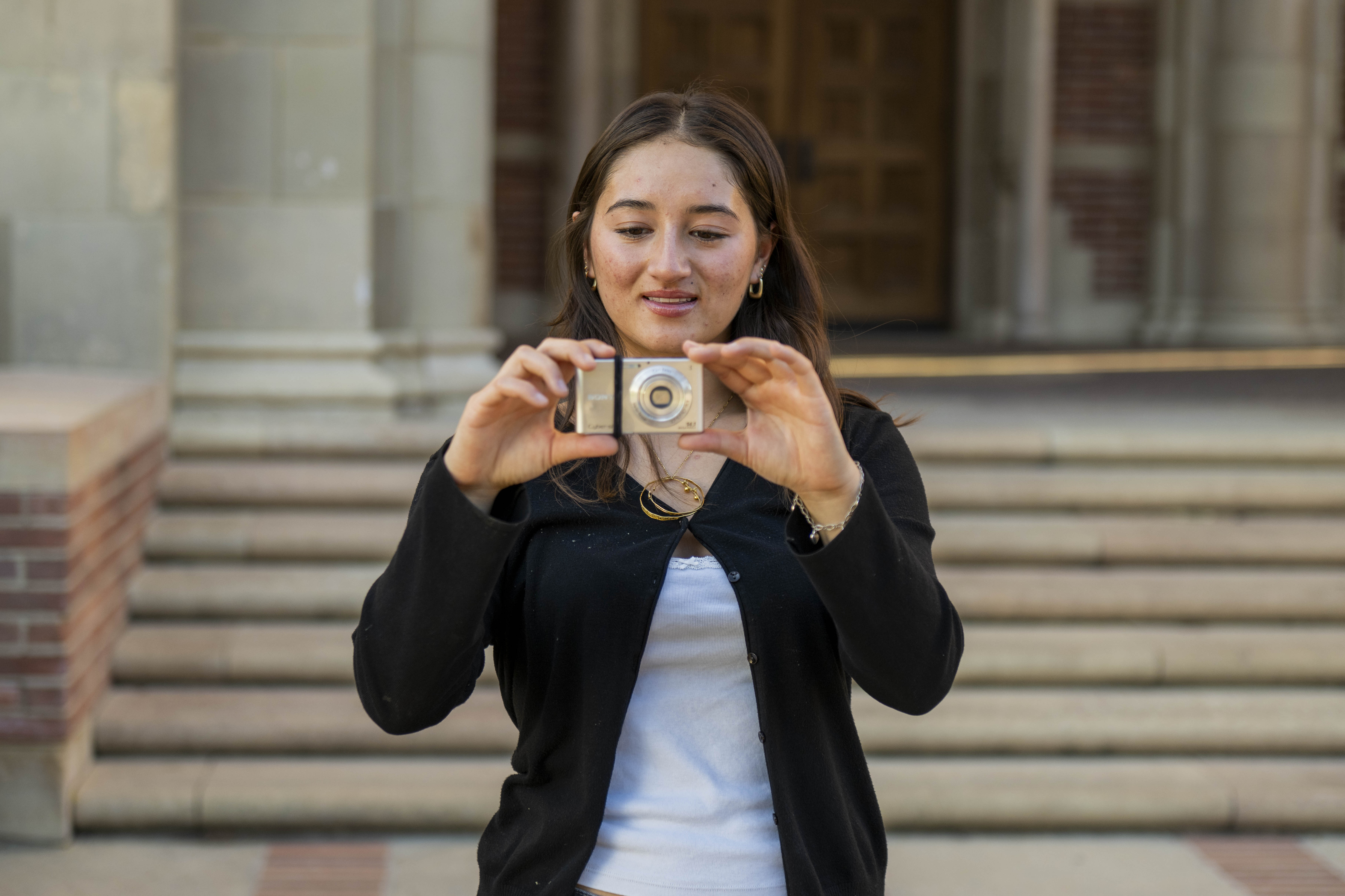 An image of a person taking a photo with a small silver camera on campus.